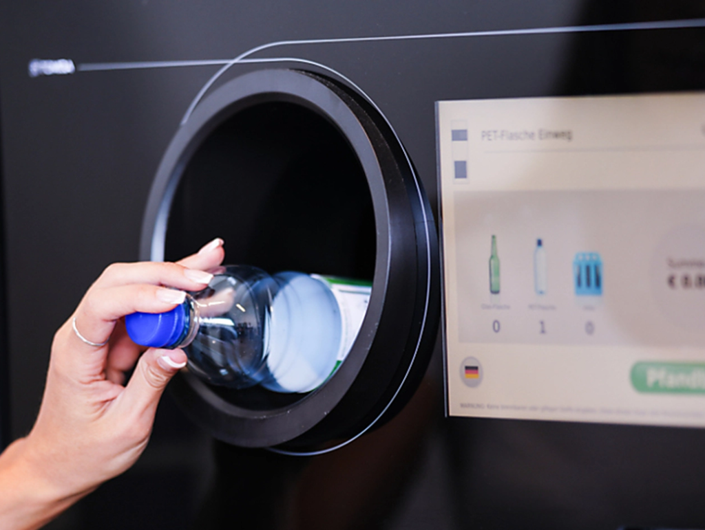 Mão inserindo garrafa plástica transparente com tampa azul em máquina de logística reversa preta com tela digital exibindo ícones de reciclagem